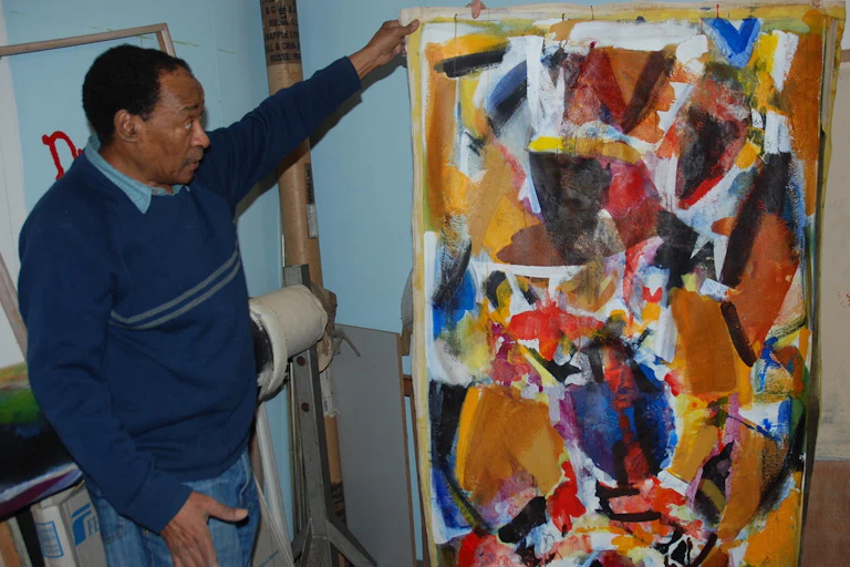 A man dressed in a navy jersey and jeans looks earnestly at a large canvas that he holds with his left arm, draped on a frame. It shows an abstract artwork of reds, oranges and blues, shapes merging and covering the canvas.