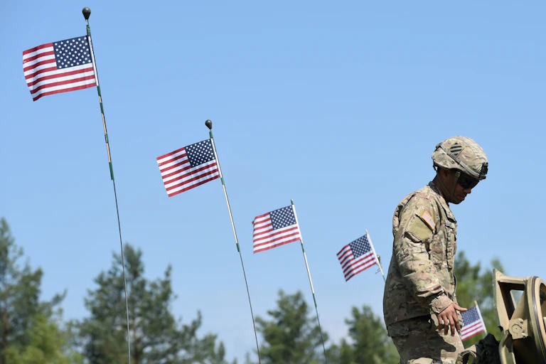 A US soldier stands at a tank with American flags flying in the background.