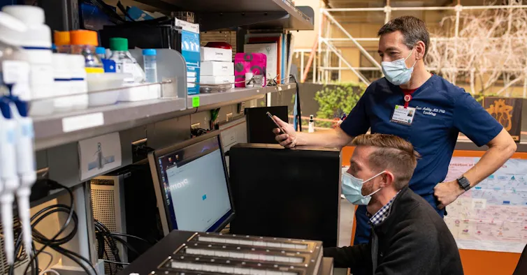 two men wearing facemasks, one seated one's standing, look at a computer screen in a laboratory