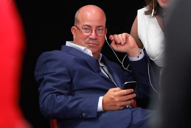 Jeff Zucker sits and stares ahead with a white earphone cord dangling from his left ear