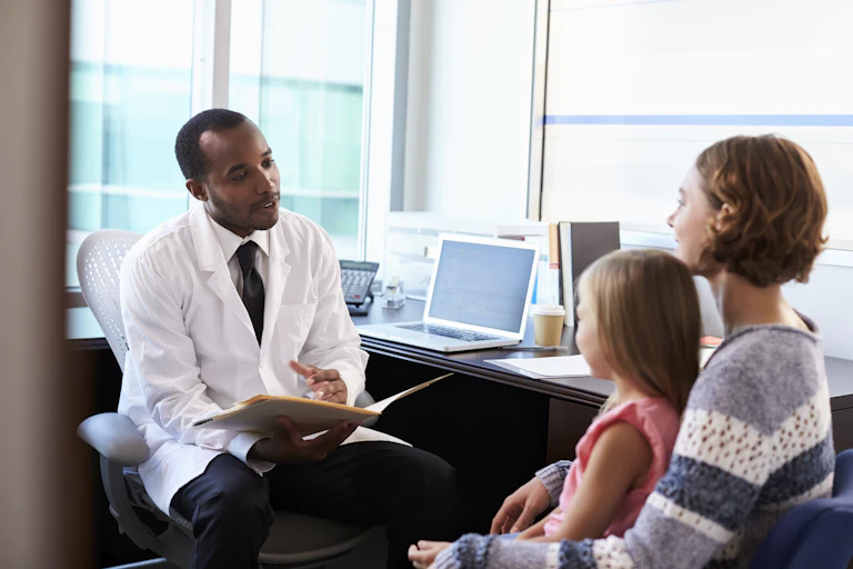 a man in a white lab coat speaks while holding an open file folder as a woman and a girl sitting in her lap look on