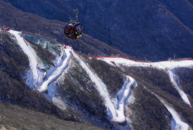 A gondola moves up a brown mountain, with a thin zigzag of snow in the background.