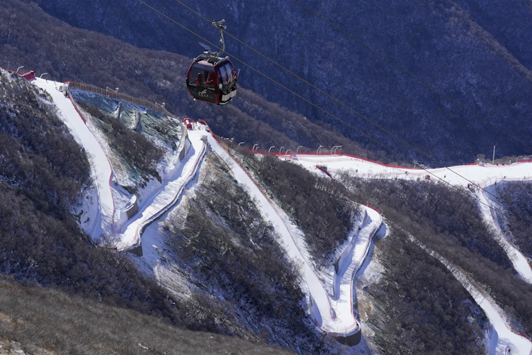 A gondola moves up a brown mountain, with a thin zigzag of snow in the background.