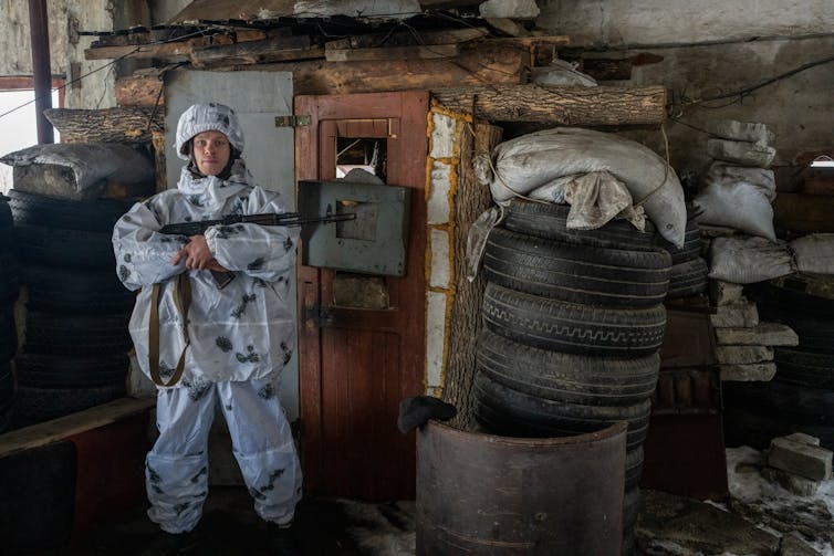 A Ukrainian solider in blue is pictured outside of a dilapidated building, with his gun across his chest