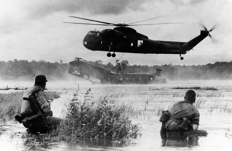 Two soldiers face a helicopter retrieving another helicopter from a body of water in Vietnam in this black and white photo