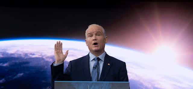 A bald man in a dark suit stands in front of a photo of planet Earth, his right hand raised.