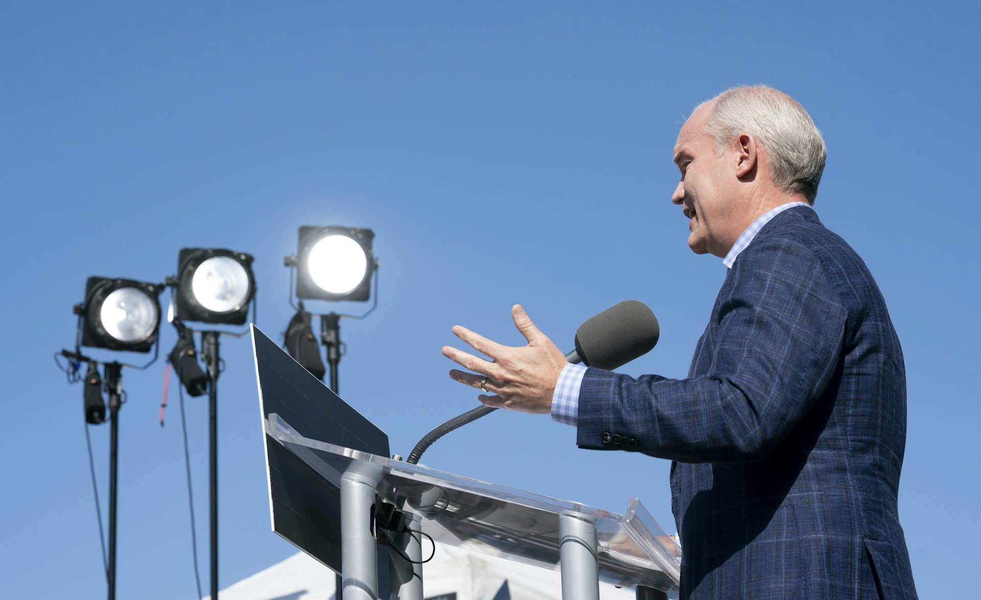 A bald man in a dark suit speaks into a microphone with stadium lights beside him.