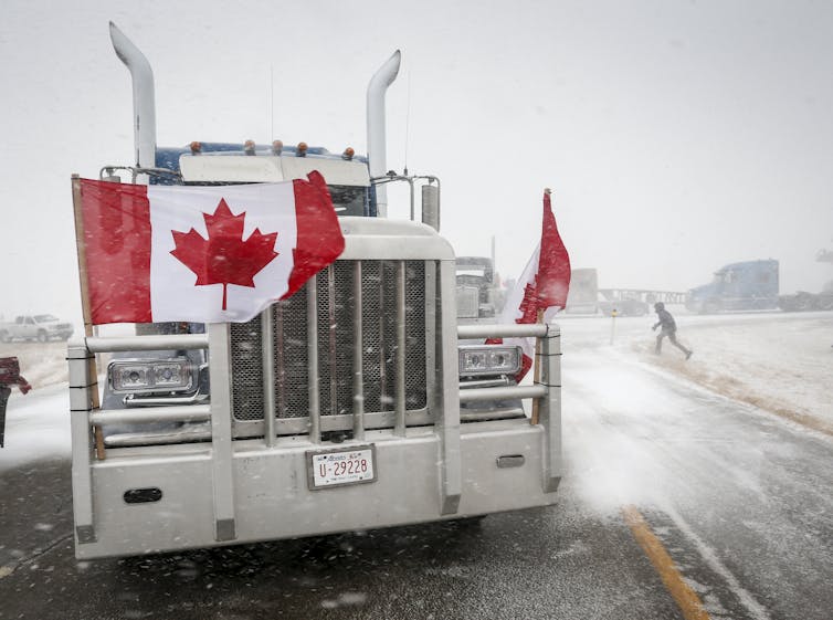 The front of a large truck on a snowy road with a Canadian blowing across its grill.