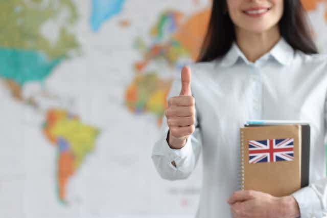 Profesora con cuaderno con bandera británica levanta el pulgar con mapa mundi de fondo.