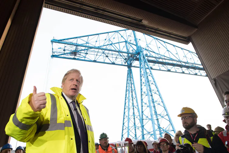 Boris Johnson wears hi-vis jacket, standing in front of a crowd of workers. A crane is visible in the distance.