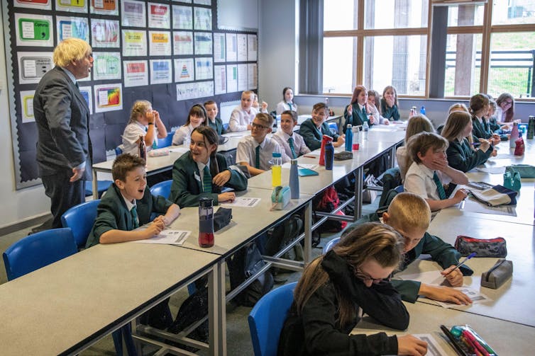 Boris Johnson stands at the back of a classroom, in which pupils sit and work.