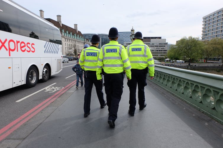View from behind of three Met police officers walking side by side across a bridge, all wearing high-vis Met police jackets