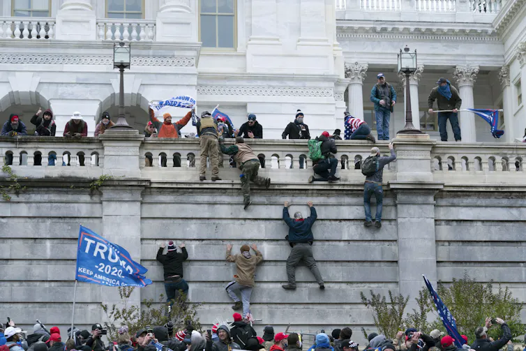 People climb the wall of the U.S. Capitol building. A blue Trump banner hangs beside one of the climbers.