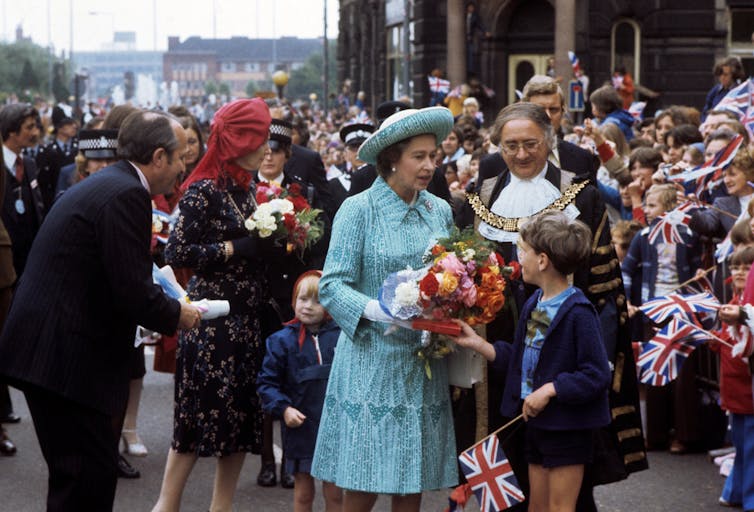 A crowd of people on a street pavement with the Queen, a mayor wearing a gold chain and a child in shorts.