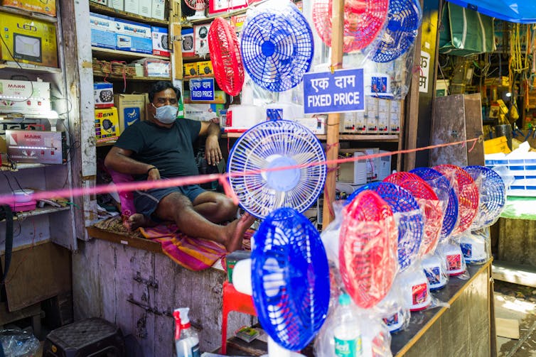 A man in shorts and a T-shirt sits in a store selling electric fans.