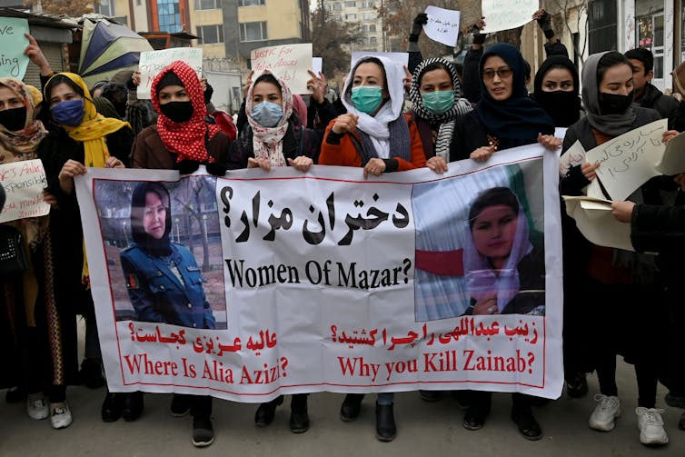 A row of women wearing headscarves and masks march in a line, holding a sign that says 'Where are the women of Mazar?'