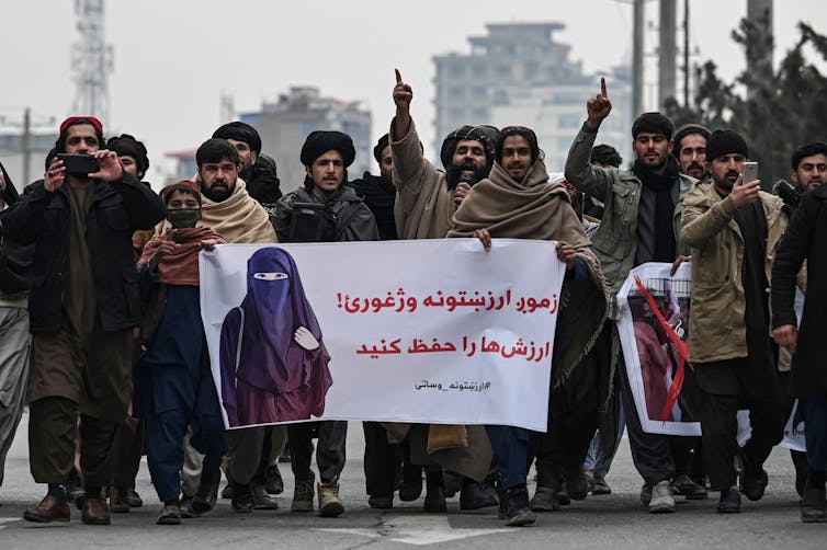 A row of men march together, holding a sign that shows a woman wearing a burqa.