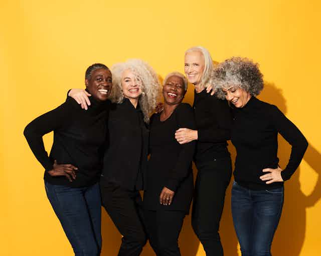 Five women of different races, all in black tops with jeans, smiling and laughing with arms around one another.