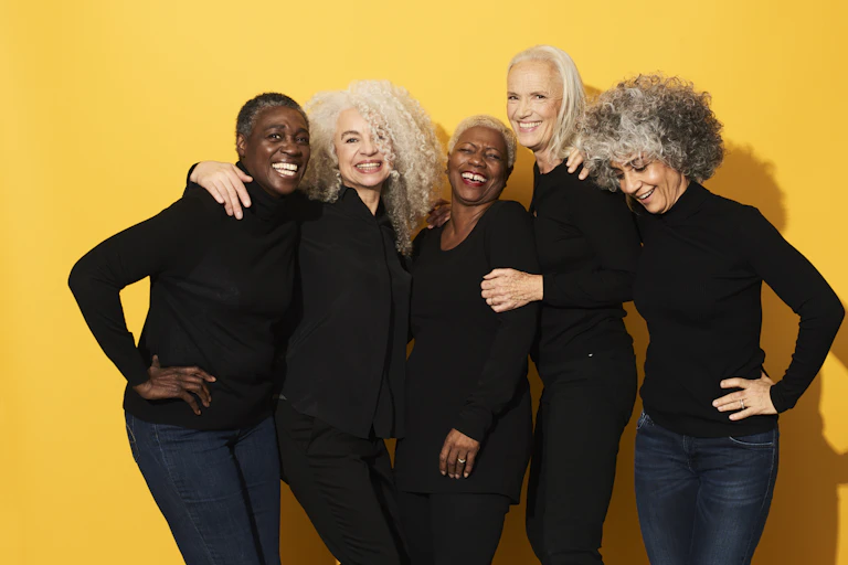 Five women of different races, all in black tops with jeans, smiling and laughing with arms around one another.