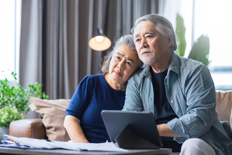 An older couple looks off into the distance, sad. They are looking at a bunch of papers on the table.