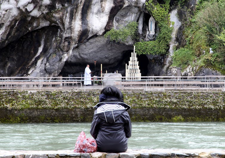 A woman with her back to the camera sits on the bank of a stream, facing a Catholic shrine.