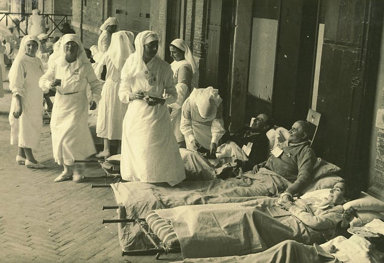 A black and white vintage photograph shows nurses in white assisting people on stretchers.