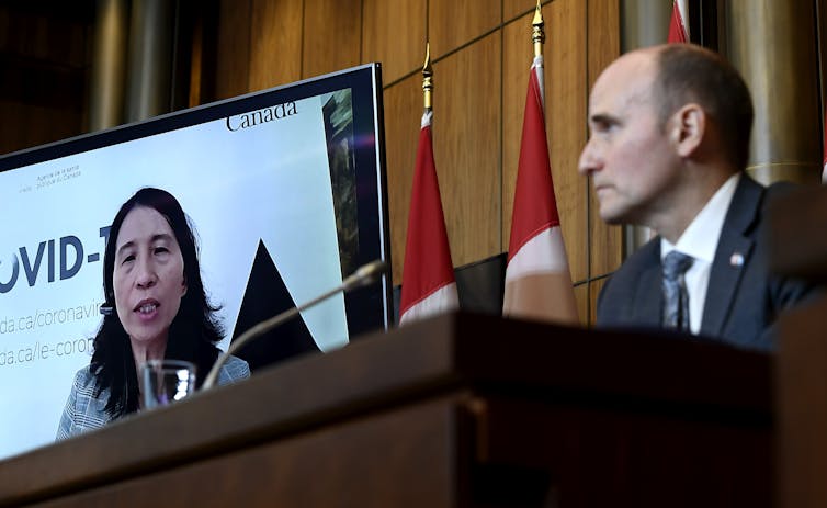 Health Minister Jean-Yves Duclos in the foreground looking at an image of Chief Public Health Officer Theresa Tam on a video screen