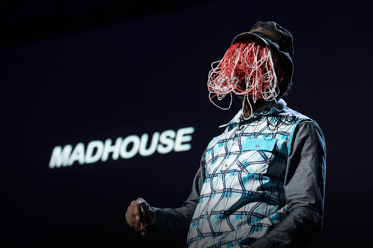A man wearing a hat and a face covering speaks to an audience