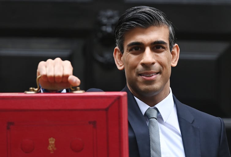 Chancellor Rishi Sunak stands outside Downing Street holding the red budget box out in front of him and looks into the camera