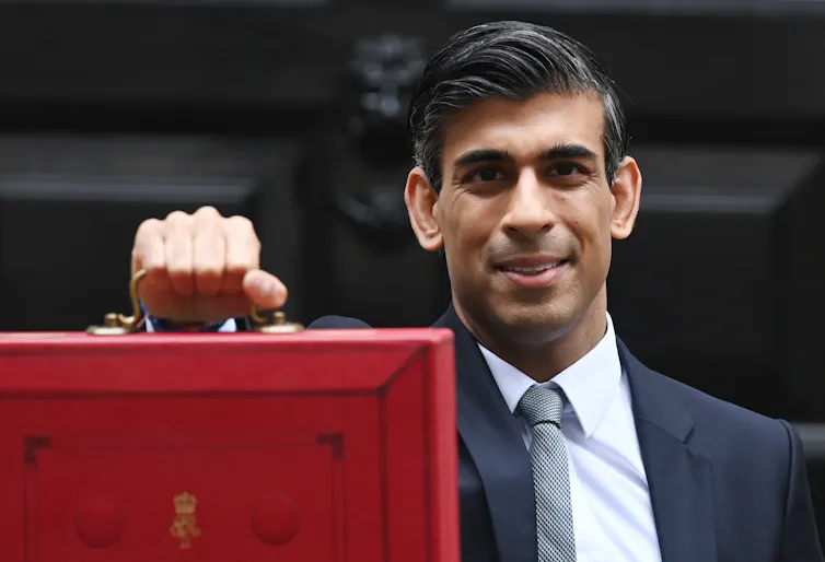 Chancellor Rishi Sunak stands outside Downing Street holding the red budget box out in front of him and looks into the camera