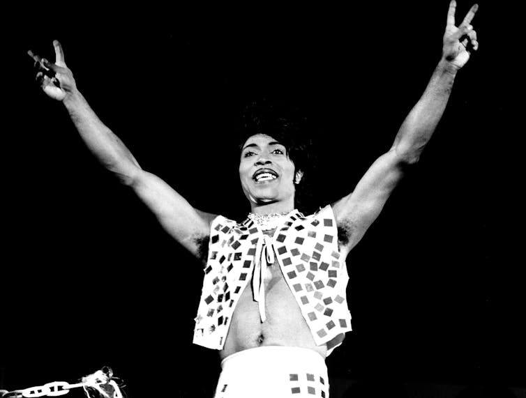 Little Richard raises arms to give two peach signs.