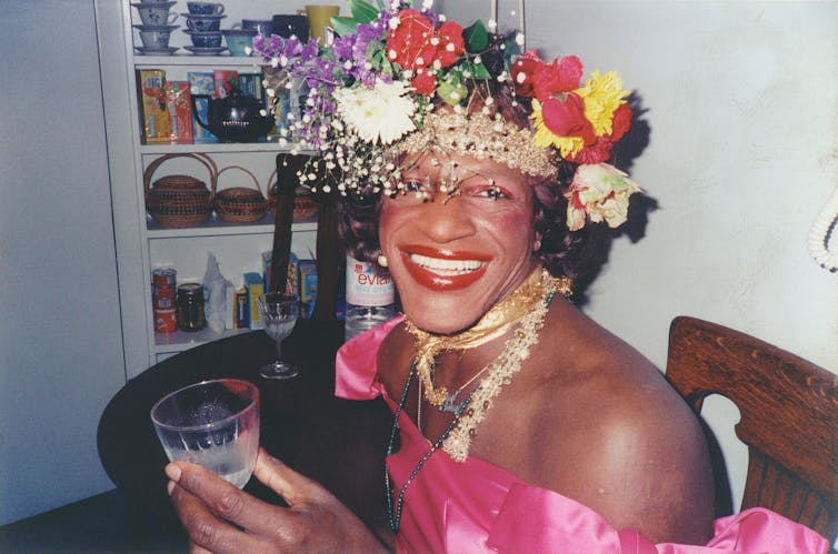 Marsha P. Johnson wearing bright clothes holding glass.