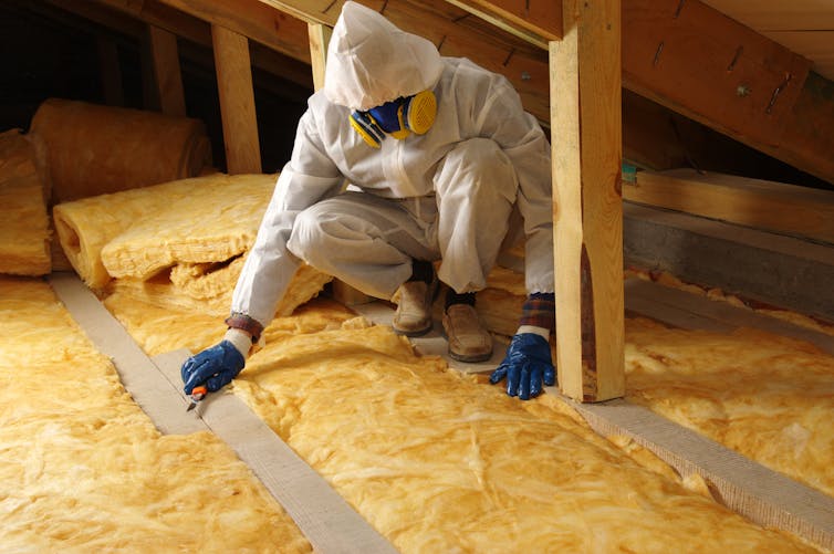 A construction worker in overalls smooths out yellow loft insulation under wooden eaves.