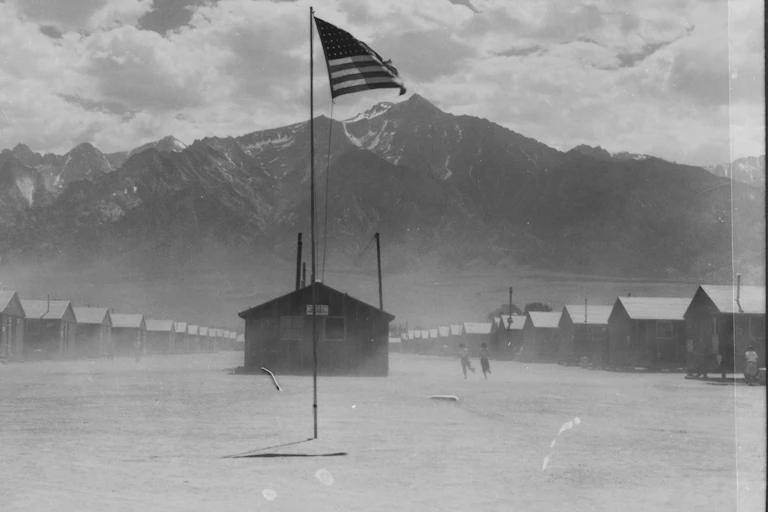 Dust swirls amid a cluster of huts, with mountains in the background and an American flag flying in the center.