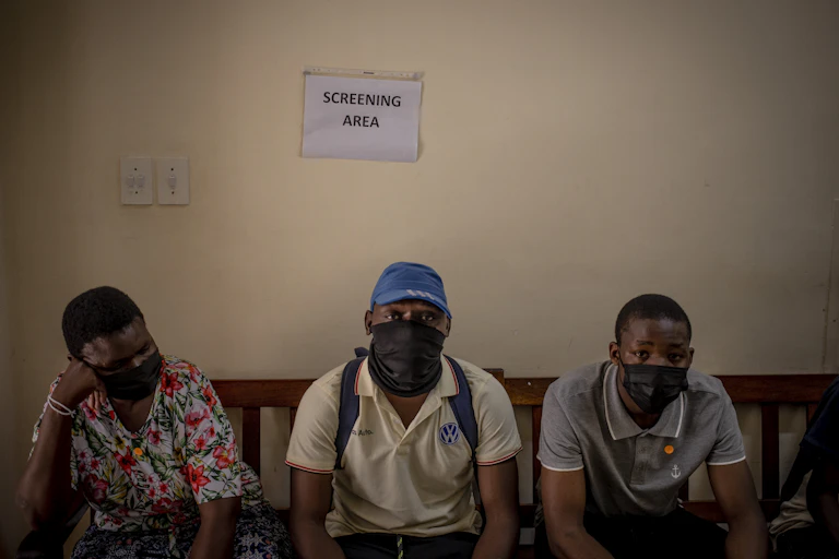 People sitting on bench with masks on with a sign saying 'screening area' behind them.