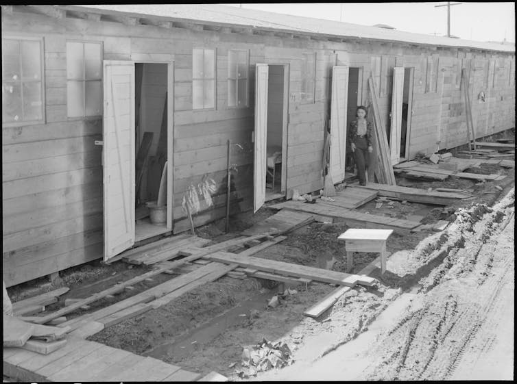 A black-and-white photo shows a woman standing in the doorway of one of a series of connected huts bordered by a muddy ditch.