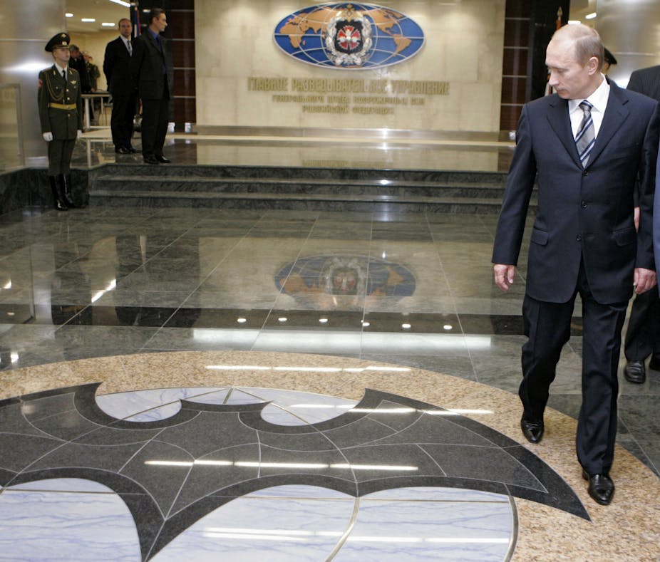 a man in a dark business suit walks through a hall with a marble floor, floor tiles in the foreground form a silhouette of a bat, an ornate crest overlays a map of the world on the back wall