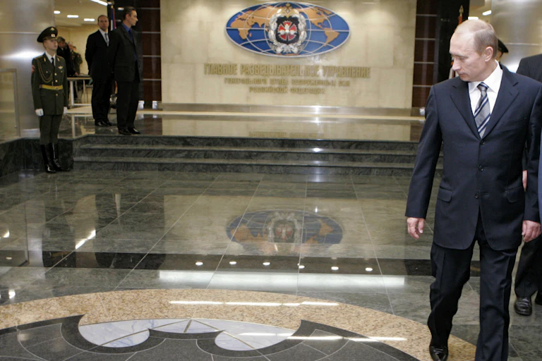a man in a dark business suit walks through a hall with a marble floor, floor tiles in the foreground form a silhouette of a bat, an ornate crest overlays a map of the world on the back wall