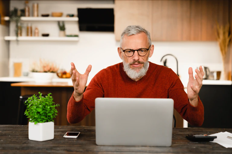 A man sitting at his laptop throws his hands up in frustration.