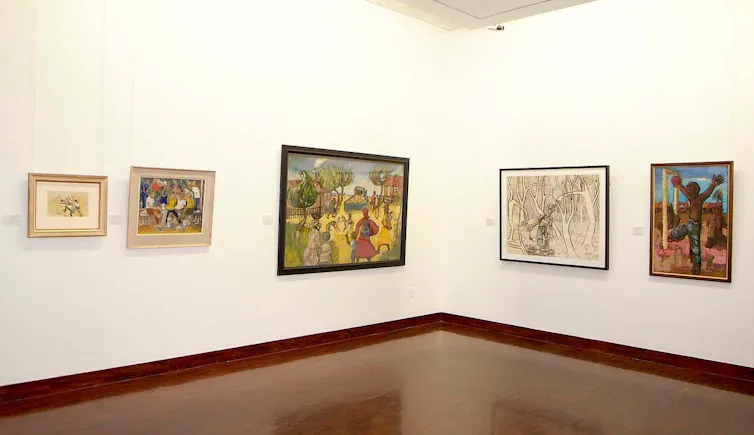 Paintings on a wall in a gallery show scenes from township life and human figures at play, in a forest, around a table.