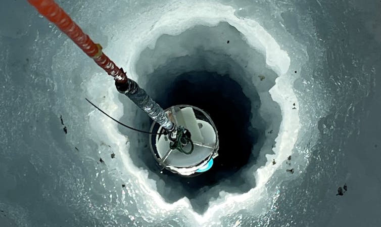 An image of camera equipment being lowered down a hole in the ice shelf. The camera is lighting up the walls of the hole, showing complex corrugations in the ice.