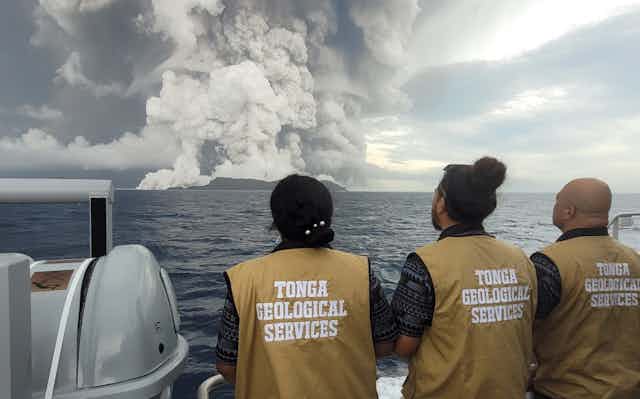 Three people wearing beige vests that read Tonga Geological Services on a boat look at smoke plumes on the horizon.