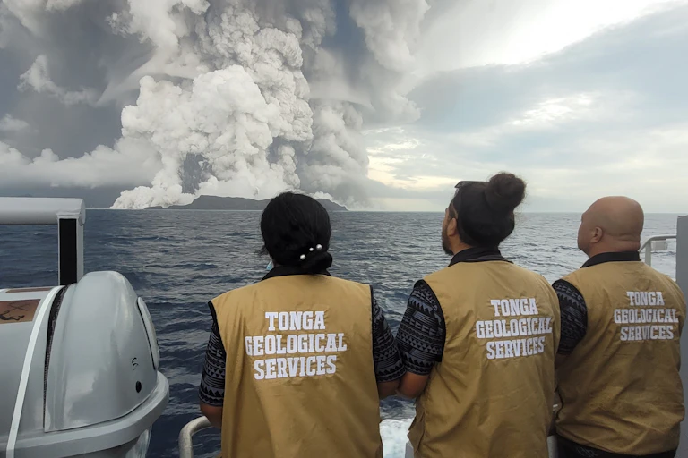 Three people wearing beige vests that read Tonga Geological Services on a boat look at smoke plumes on the horizon.