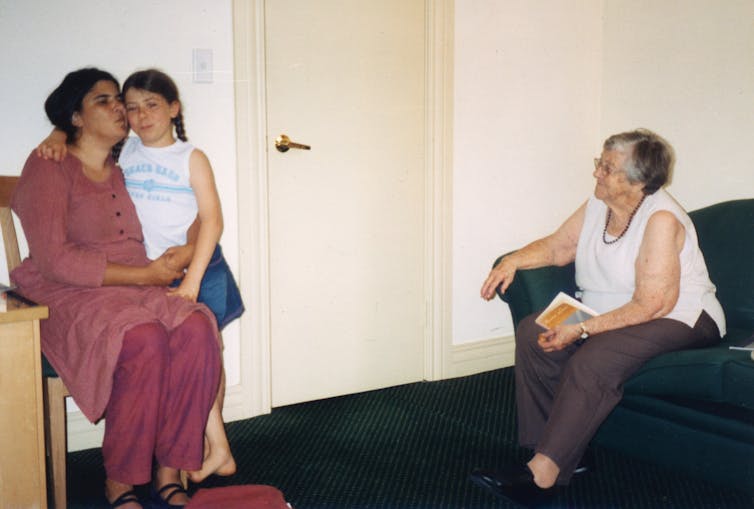 A young girl embraces her seated mother, who kisses her cheek, as an older woman looks on nearby.