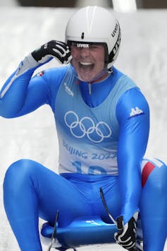 Luge athlete Dominik Fischnaller of Italy dressed in blue with a white helmet, smiling as he reaches the bottom of the track at the 2022 Winter Olympics.