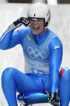 Luge athlete Dominik Fischnaller of Italy dressed in blue with a white helmet, smiling as he reaches the bottom of the track at the 2022 Winter Olympics.