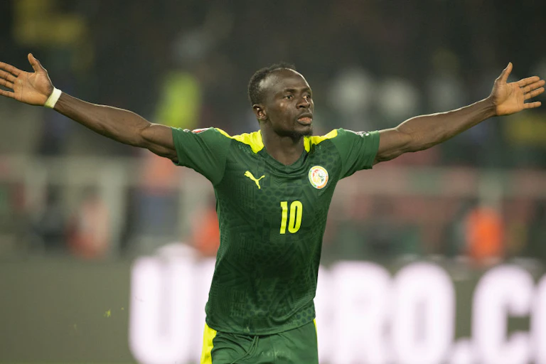 A ma in football uniform of green and gol and bearing a number 10 on the chest runs on a field, both his arms stretched out in victory.