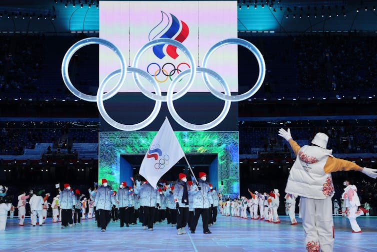 People wearing cold-weather clothes parade through a stadium under the Olympic Rings
