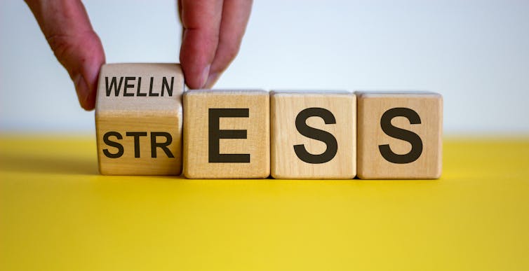 Four blocks with letters on them. A person's hand is turning one block over, so that the word the blocks spell can read either 'wellness' or 'stress'.