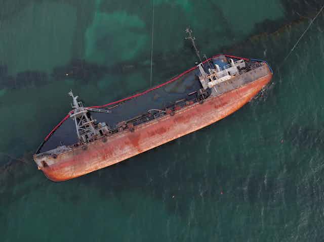 A grounded ship rests on its side and leaks oil into the ocean.